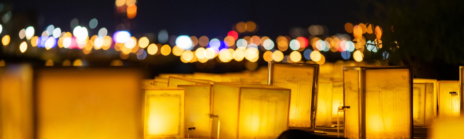 Obon paper lanters float on a stream under distant colored lights.