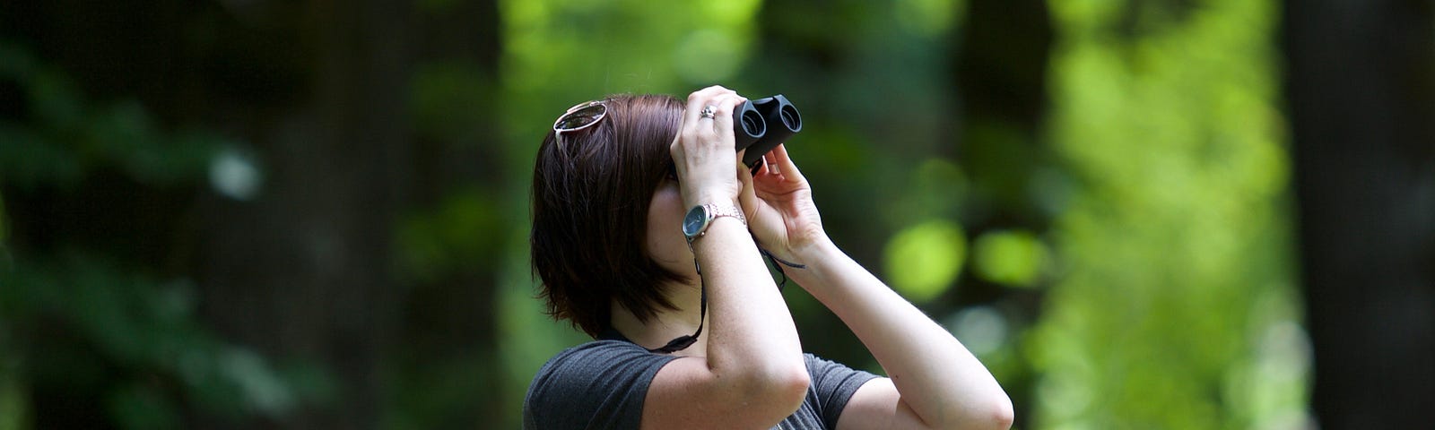 Woman in a forest looking up at trees with binoculars.