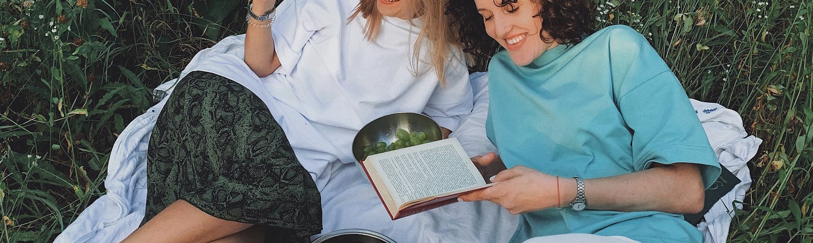 Two women sat on grass reading a book and eating olives