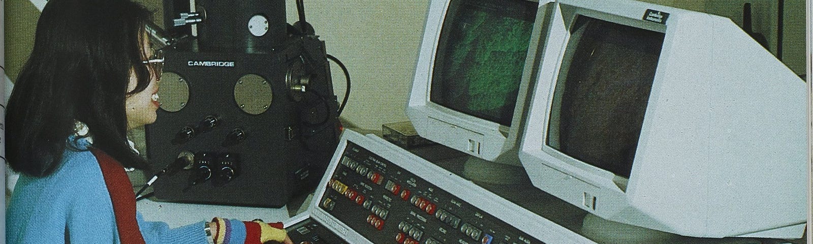 A student in striped fingerless gloves, a blue jumper and glasses sits at a desk at the keyboard of a large electronic machine with several rows of coloured buttons and two white monitors.