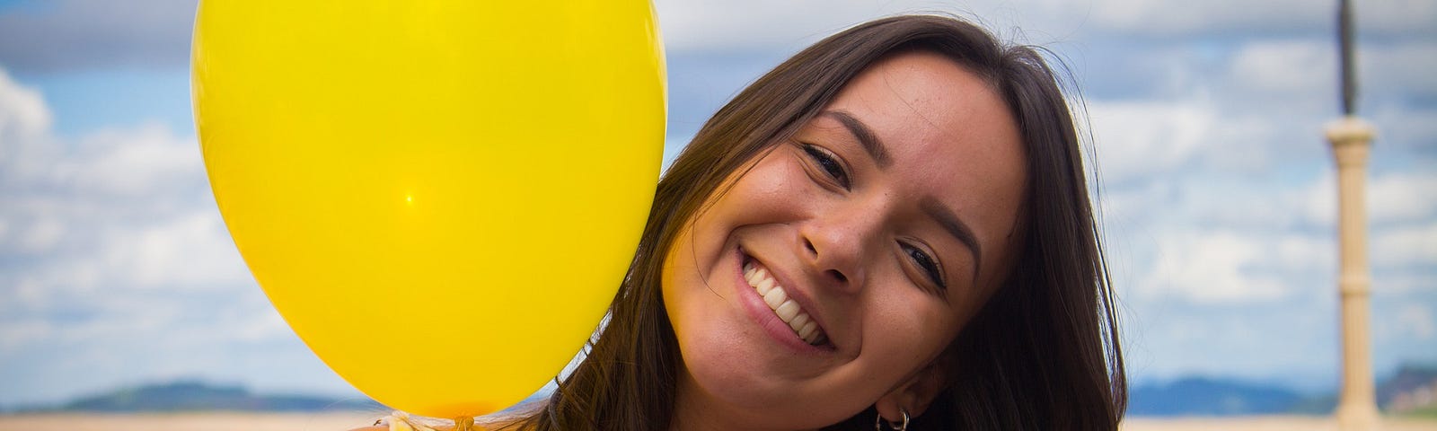 Young woman with long dark hair smiling with her head tilted to her left. On her right, by her face, is a yellow balloon.