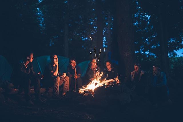 A group of people around a camp fire. Tents and woods in the background/