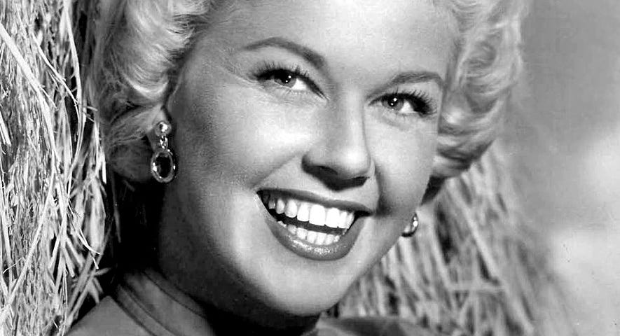 Doris Day, with blond curly hair, is leaning against a bale of hay, smiling and looking directly at the camera.