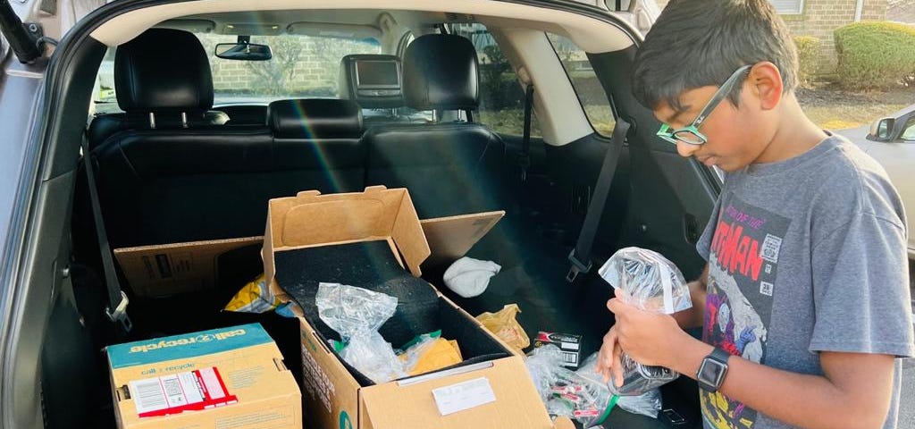 Brown boy with short black hair wearing turquoise glasses filling boxes in a trunk on a sunny day