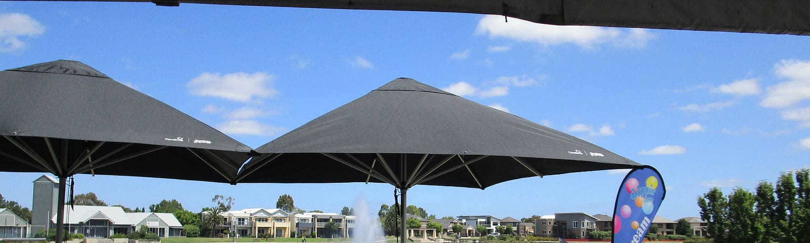 A cafe terrace in the foreground overlooking an ornamental lake & fountain with residential & community buildings beyond.