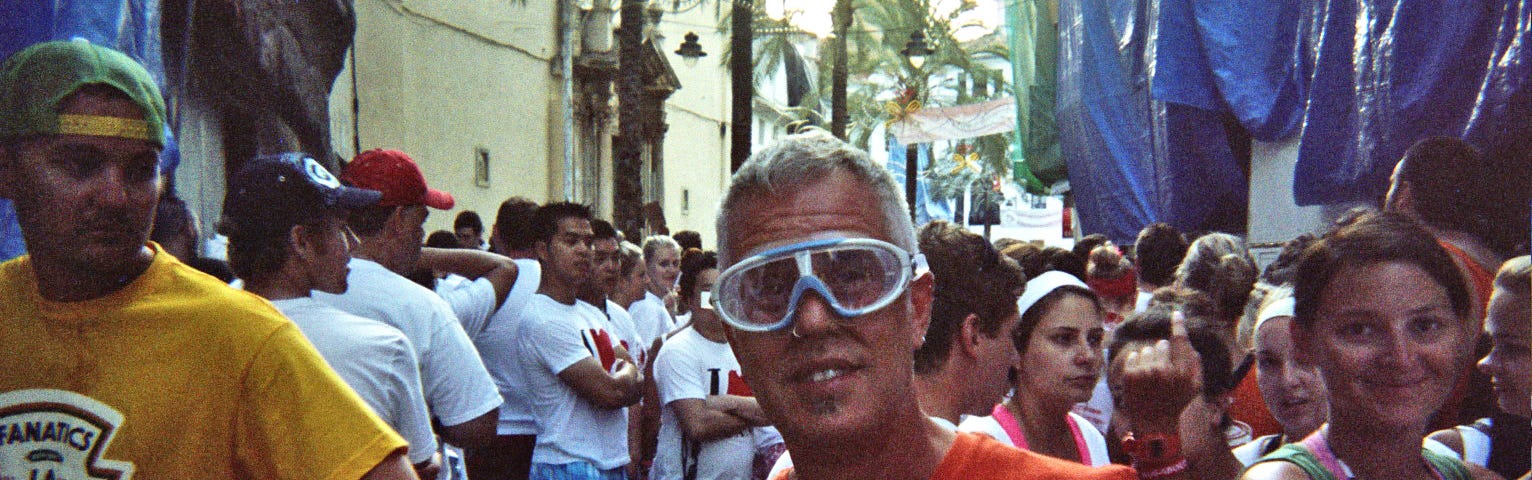 man stands in the middle of a crowded street wearing an orange t-shirt and water goggles.
