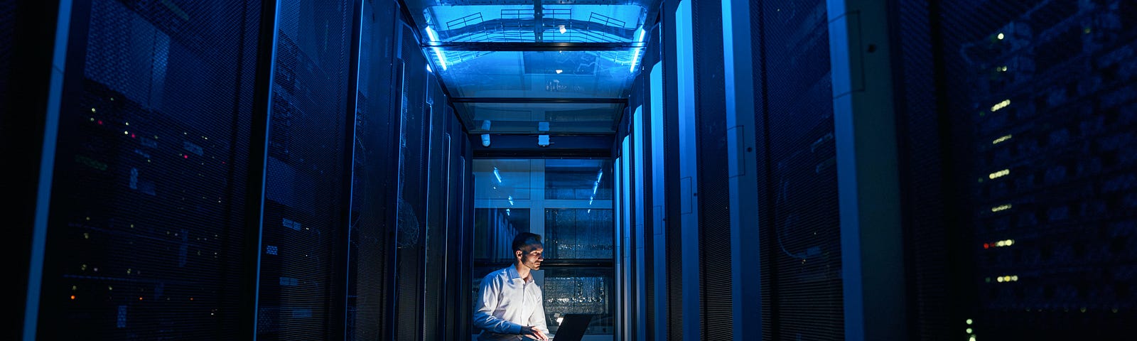 Man in dress shirt kneeling with computer in front of computers.