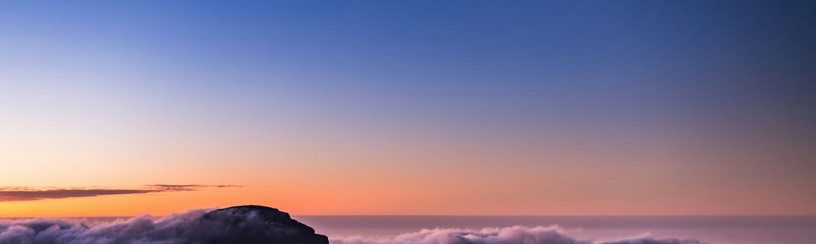 A view of dawn over a mountain top laced with clouds, the top of the photo is a deeper blue and the horizon orange and lavender
