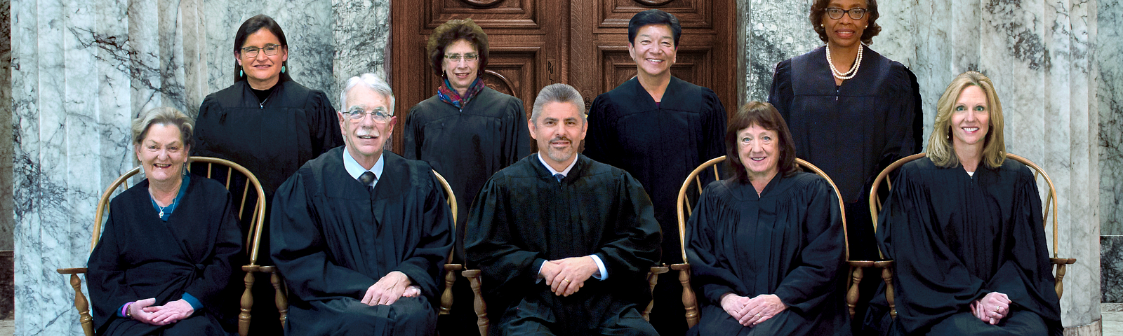 Five justices of the Washington State Supreme Court sit side by side on chairs while four more stand behind them. They are wearing their black robes and are positioned in front of the ornate wooden doors of the court room at the Temple of Justice.