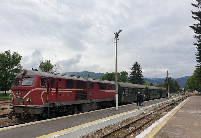 red train engine with green carriages sitting at quiet train platform under cloudy skies