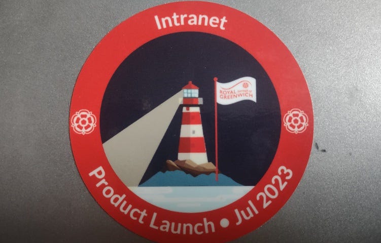 Close up on a sticker on a laptop lid. The sticker reads ‘Intranet Product Launch. Jul 2023’. The design on the sticker is a striped red and white lighthouse on a rock shining a light onto the sea. There is a flag with the Royal Borough of Greenwich logo on it next to the lighthouse, and two tudor rose emblems (also part of the borough’s logo) on either side of the sticker.