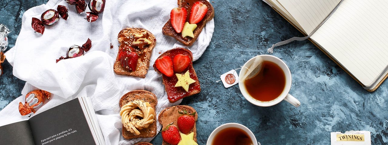 Two open books surrounded by cups of tea, chocolates and toast with delicious strawberry topping.