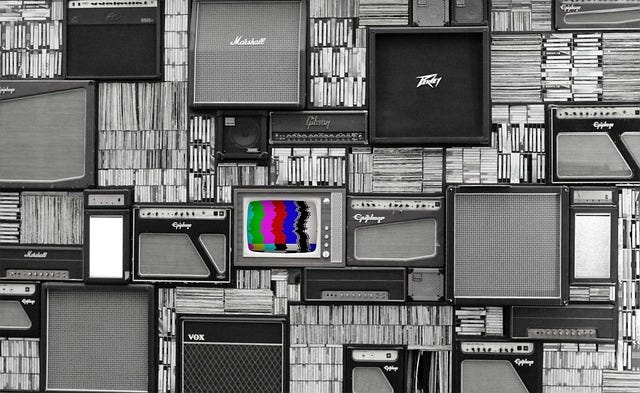 A wall of amplifiers with a TV in the middle. The amplifiers are in black and whtie but the TV has colored squiggly lines.