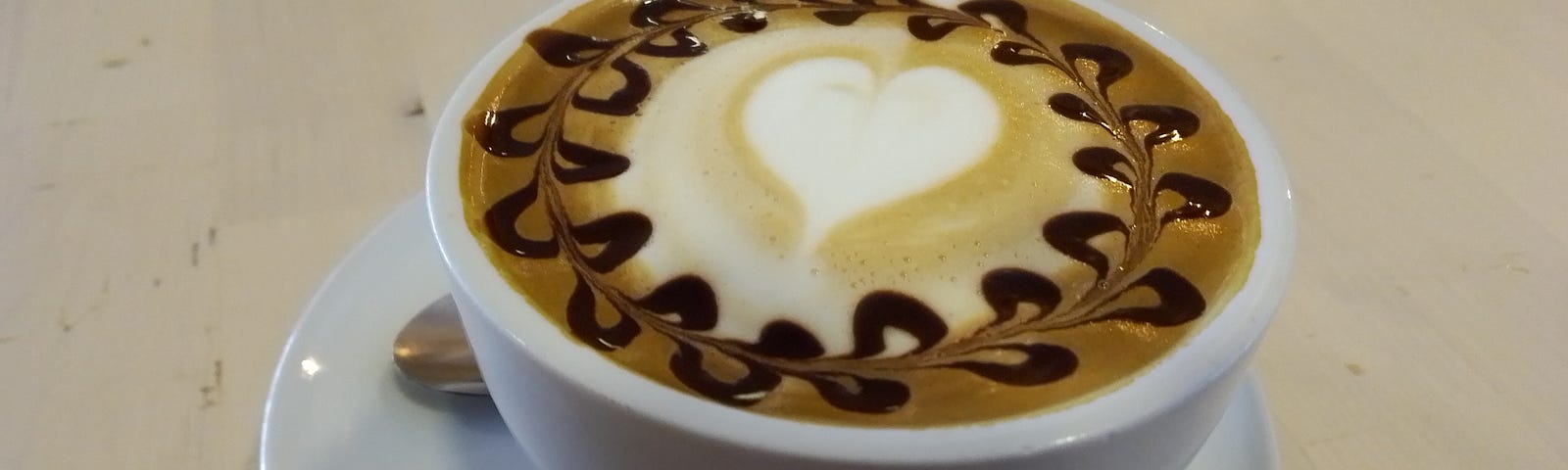 Cappuccino with a froth heart in the center and yummy chocolate syrup squiggles around the edges, served in a white cup.