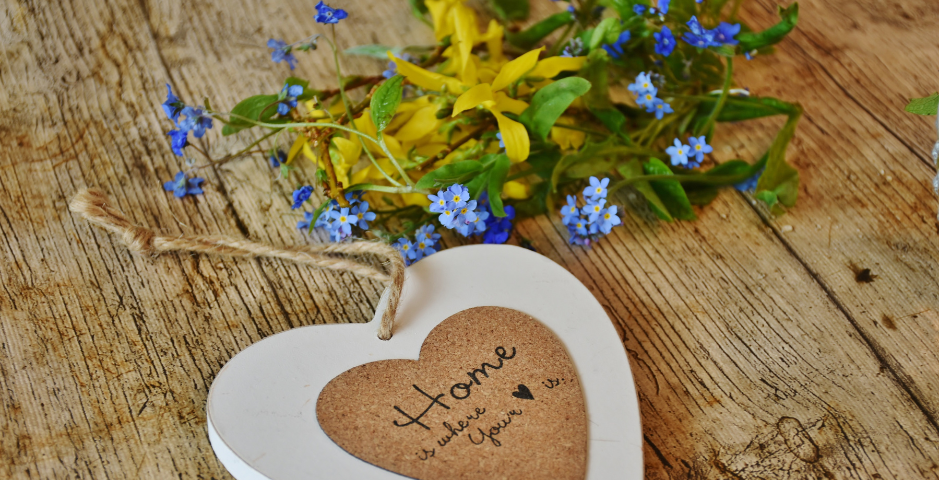 heart decor and flowers placed on a wooden table, written on the heart are the words, “home is where the heart is”