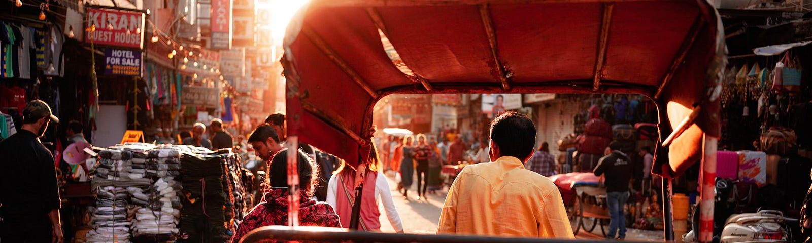 Man pulls an empty rickshaw on a street in Delhi. Shops line both sides of the road with people looking through various items.