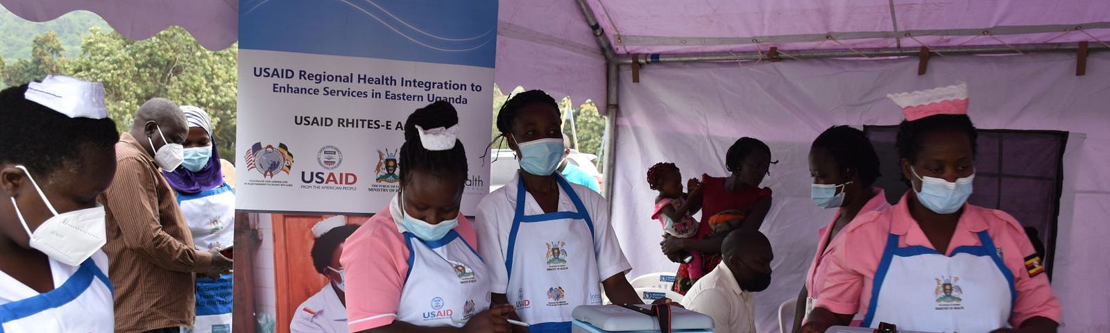 Health workers in nurses caps and masks stand under a tent covering a table filled with coolers and supplies to administer vaccinations.