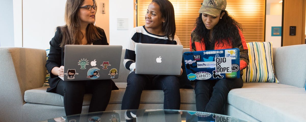 Three women of colour sat on a sofa with their laptops open with a low table in front of them