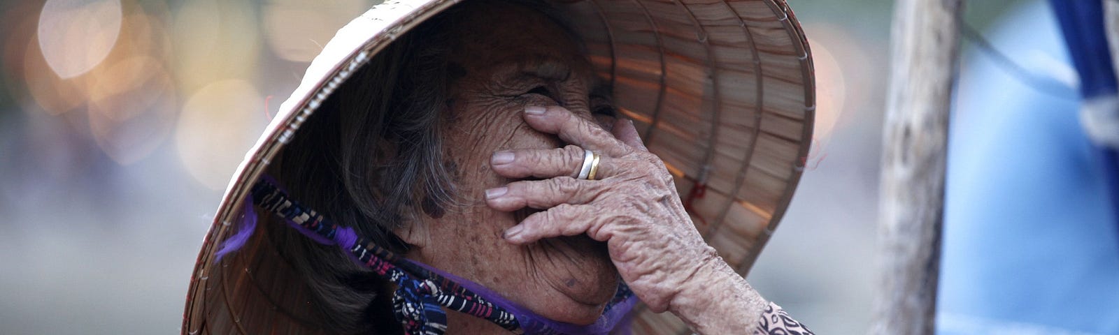 Old woman in bamboo kasa hat, laughing, covering her face with her hand.
