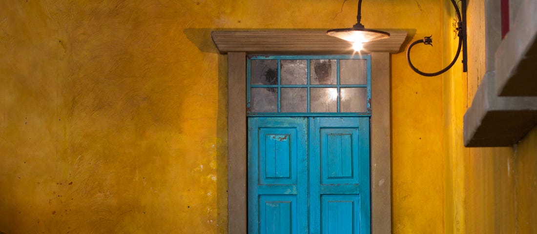 an old blue door in a yellow wall with a old lamp shining softly.