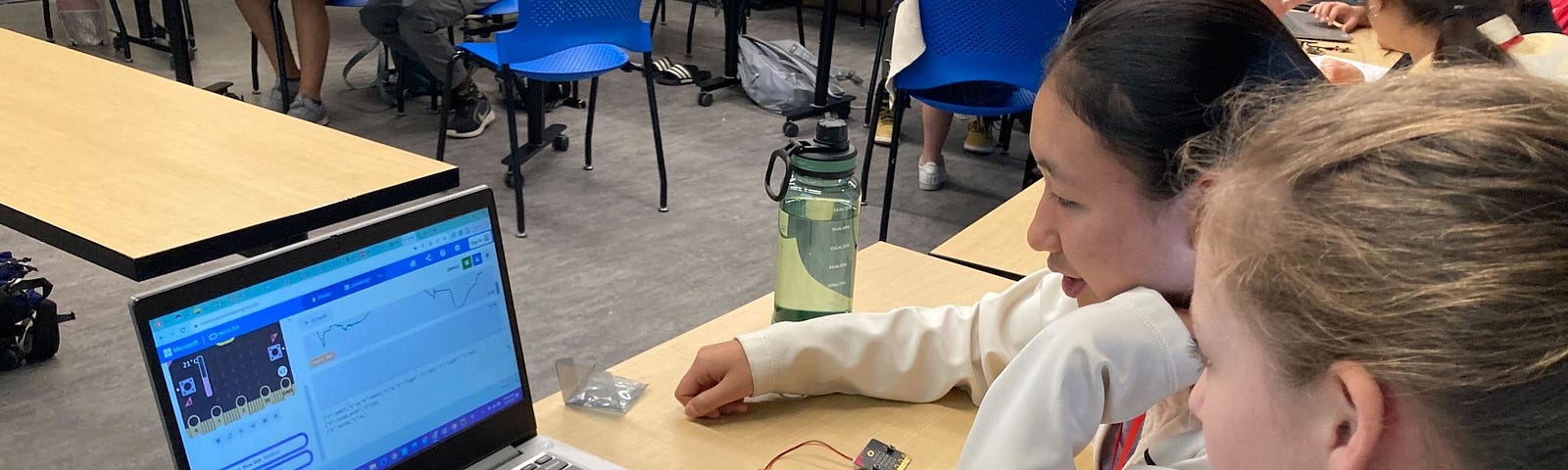 Photo of a middle school classroom with a dozen students coding on laptops at different tables. A mentor is speaking with 3 students in the back.