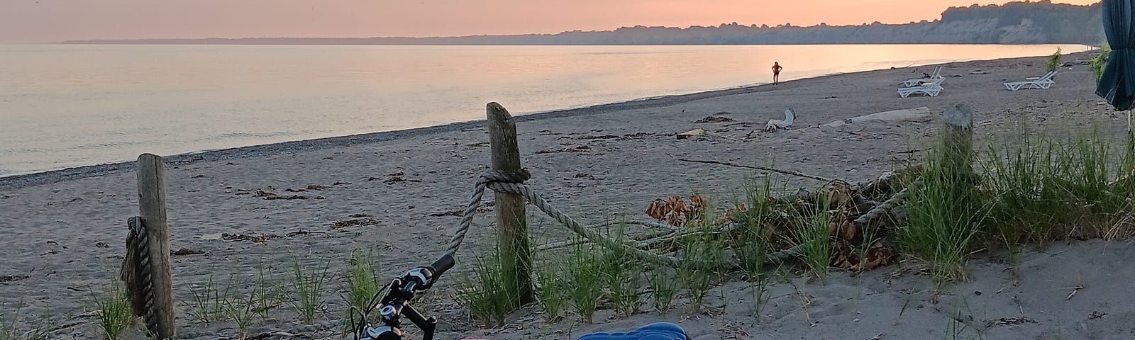 A bike leans against a picnic table that has a pink helmet and green water container on it, with a beach and lake in the background.