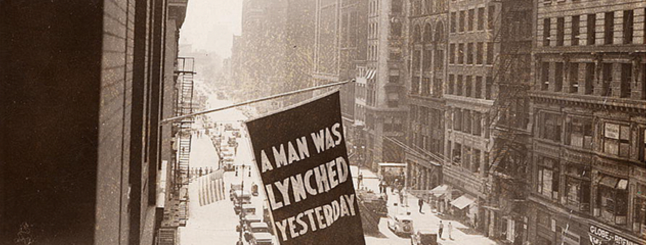 A black and white photo of a flag hanging in a city, reading “A man was lynched yesterday.”