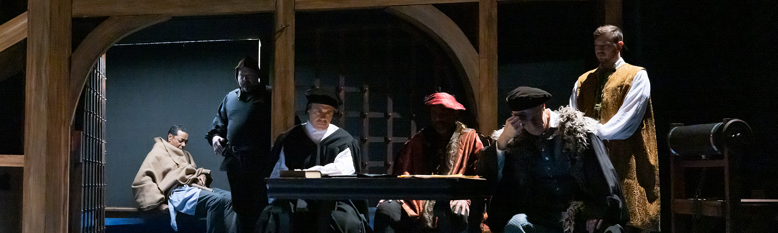 A man sits in a prison cell under a brown blanket guarded by a jailer, while four men in 16th century clothes sit at a table outside, looking frustrated.