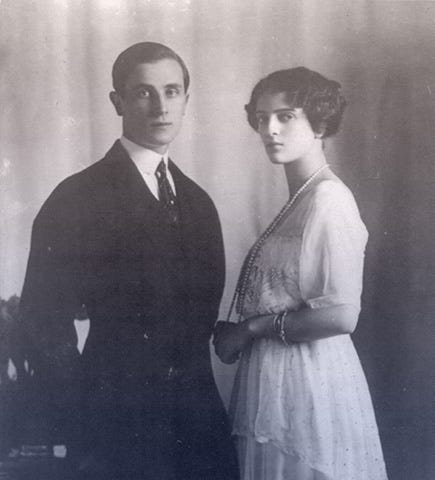 Felix and Irina standing together. He’s in a dark suit with short, slicked-back hair. She’s in a white day dress with pearls.