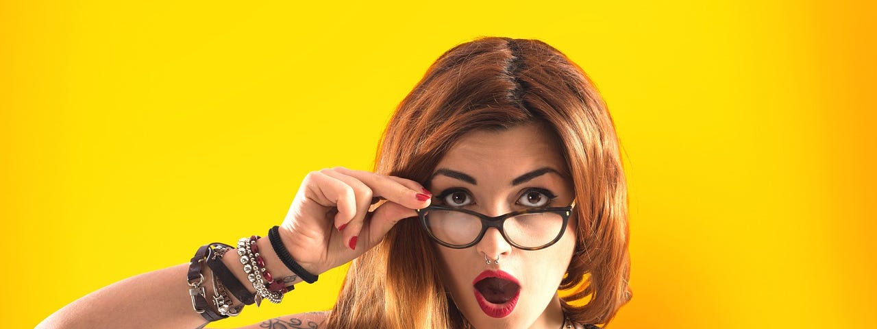 A red-haired woman wearing glasses in front of a yellow background, looking astonished