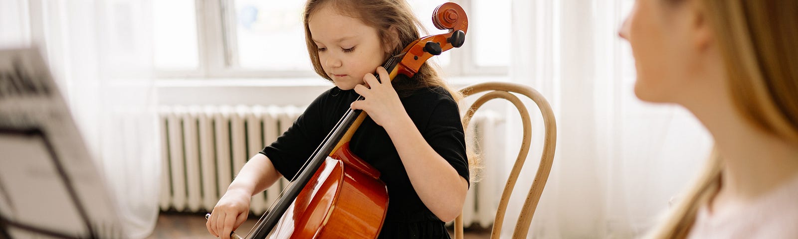 A young girl playing cello in a bright sunlit room. On the right side of the photo, an adult woman is watching her.