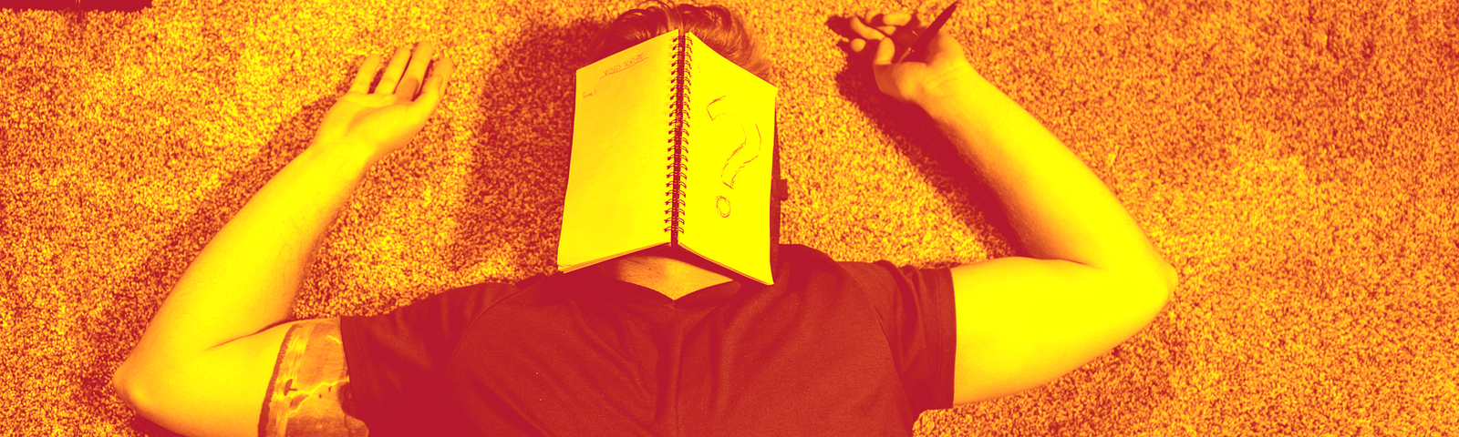 A man is laying on the floor with a notebook open over his face with a pen in his left hand. The notebook has a question mark drawn on a page using a blue pen.