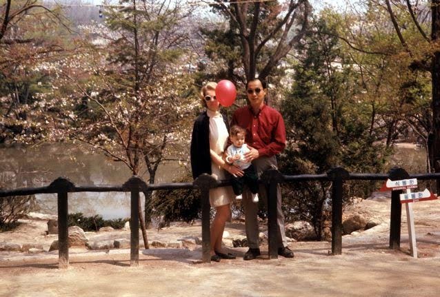 A photo from 1968 of me as an infant with my parents in a park. We’re in front of flowering trees and a pond, leaning against a wooden fence. My father is wearing a red shirt and black pants, my mother is wearing a white dress with a black coat, and a red balloon floats in front of her face. They are wearing sunglasses.