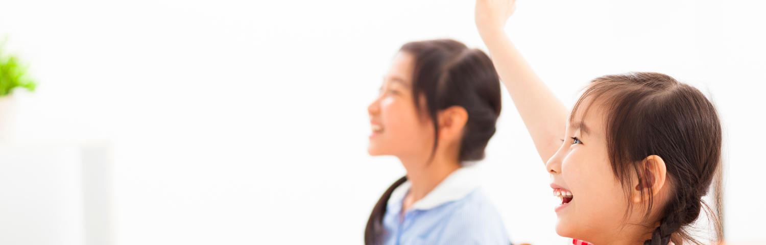 Two female students of Asian heritage sit at desks in a classroom. One raises her hand and both are smiling.