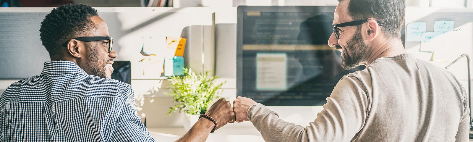 Two guys fist bumping in front of a table and computer screen