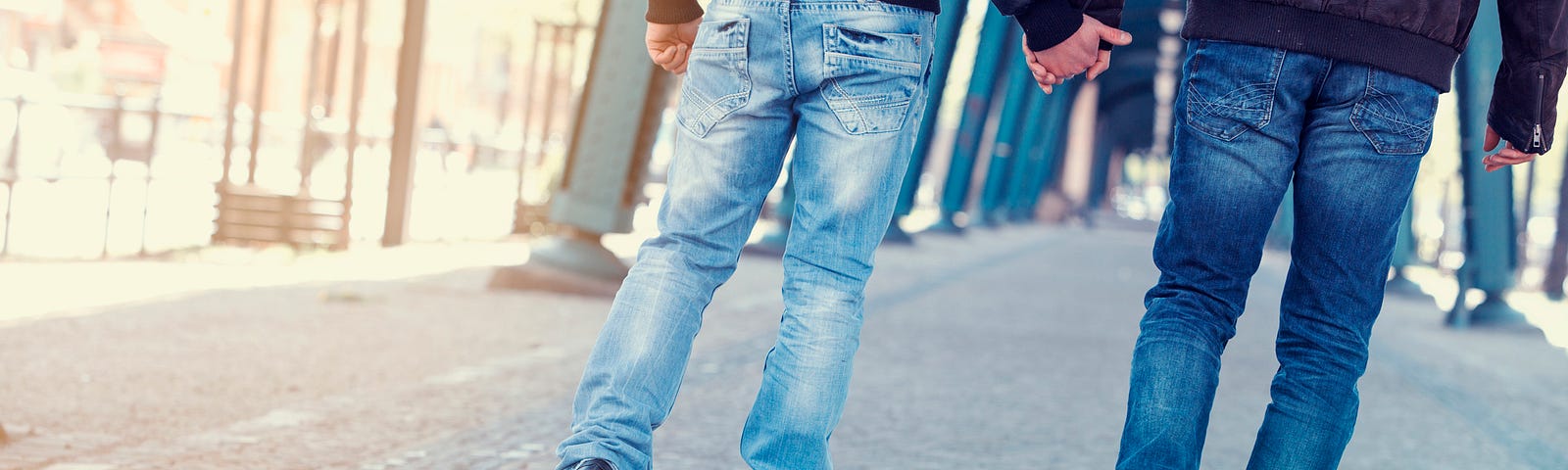 Two men in jeans, holding hands and walking. Photo from back