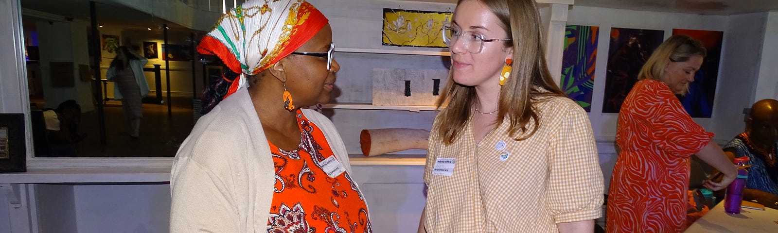 A black woman with a red dress and headscarf stands on the left, talking with a white blonde woman with a yellow top and white skirt on the right.