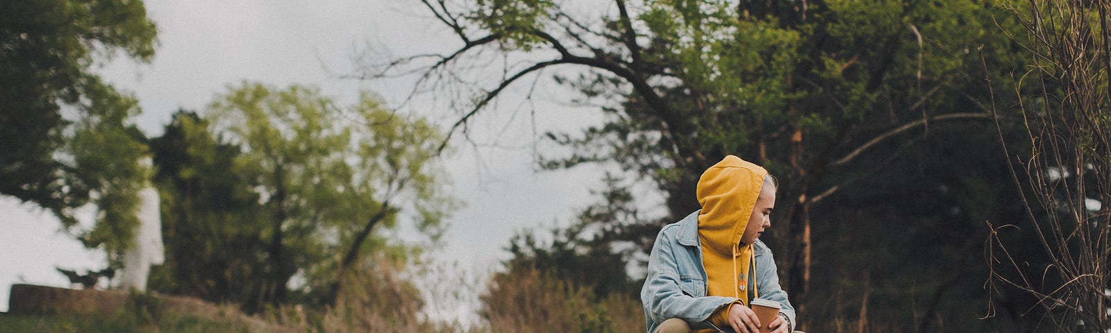 A woman in a yellow hoodie and a denim jacket sits alone on a fallen tree. She holds a cardboard-clad coffee.