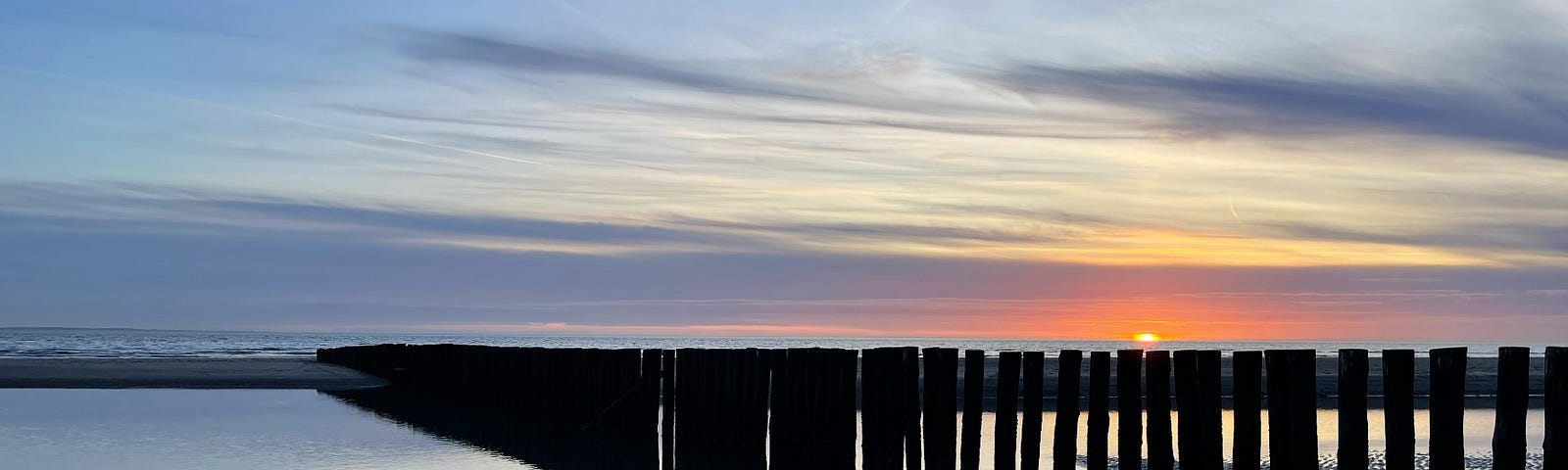 A blue-gray photo of the sunset as seen from the beach. From the right, a long row of wooden poles disappears towards the horizon. They are reflected in the quiet water of the North Sea, a tranquil and colorful photo.