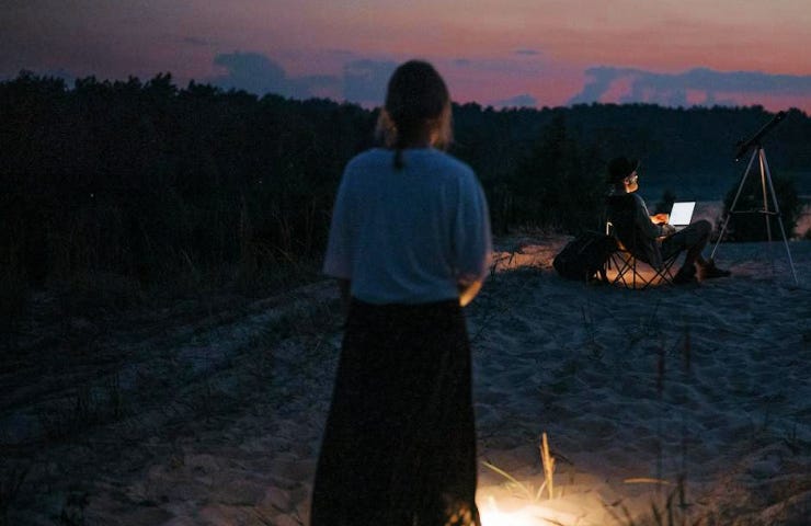 From the back, we see a woman standing on a dark beach.