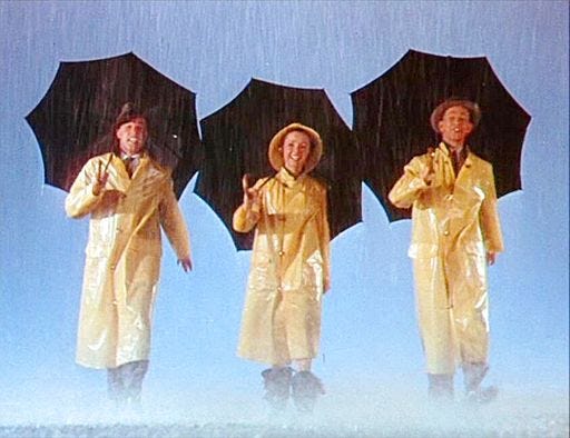 Three people side by side wearing yellow slickers and rain boots while carrying black umbrellas.