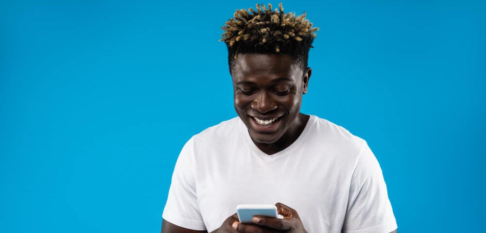 Portrait of young man in white t-shirt using mobile phone and smiling. Isolated on blue background