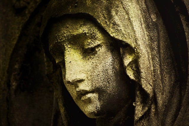 A yellowish, roughhewn stone cemetery statue of a woman’s face, dark back ground.