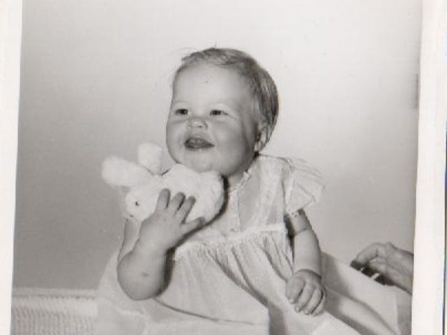 Black and white photo of a chubby cheeked 1 year old girl in a dress and holding a stuffed bunny under her chin.