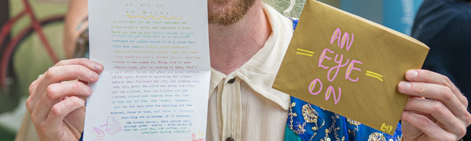 Photo of the writer Luke Winter wearing an embroidered blue jacket, sat in front of a typewriter, holding up a personalised typed story and envelope.
