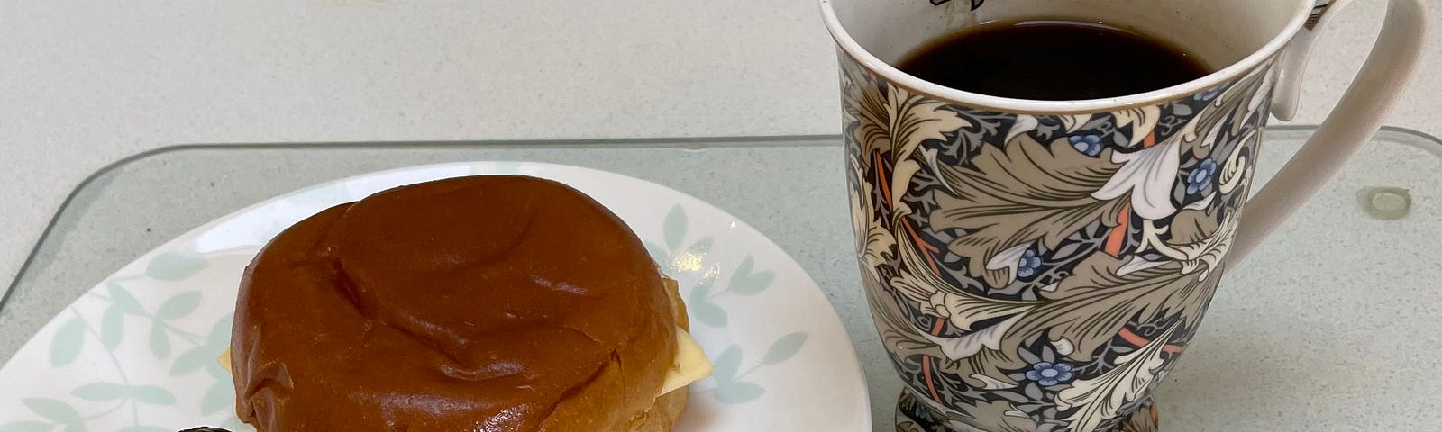 A shiny brown brioche bun with cheese poking out its edges, sits on a white plate next to two small green pickles. A mug of espresso stands next to the meal.