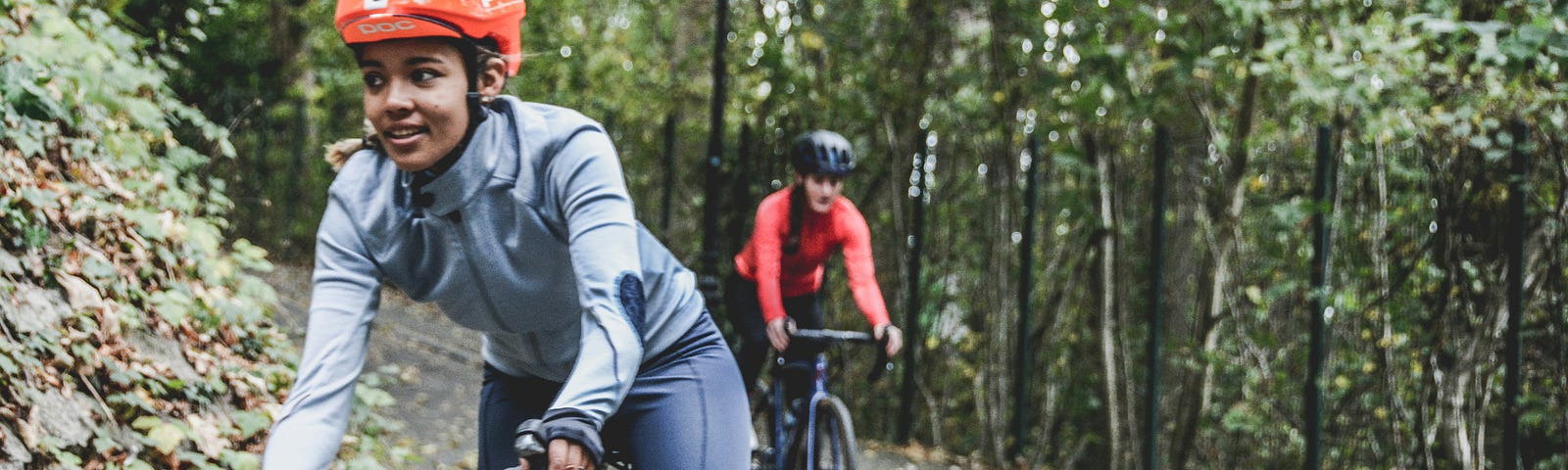 2 women on bicycles, wearing helmets. Bicycling on a path near a forest. Photo by Coen van de Broek on Unsplash