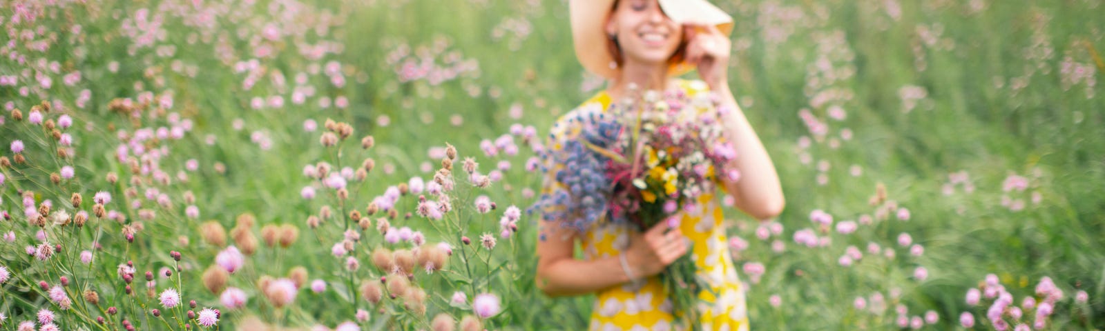 Out-of-focus girl in yellow and white floral dress standing on flower field during daytime holding a bunch of flowers.