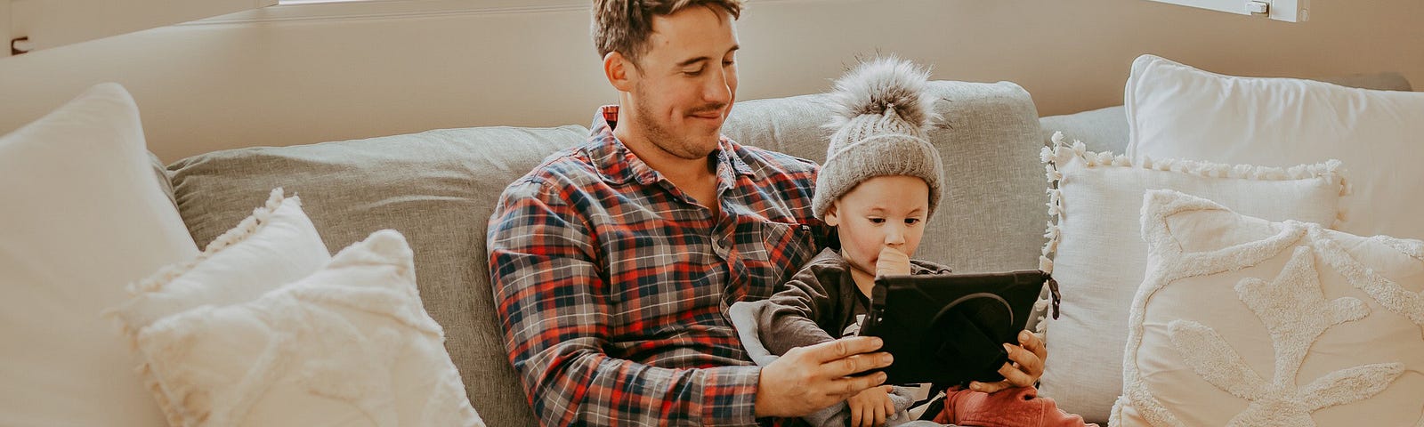 A father and young son sit on a couch watching something on an iPad together.