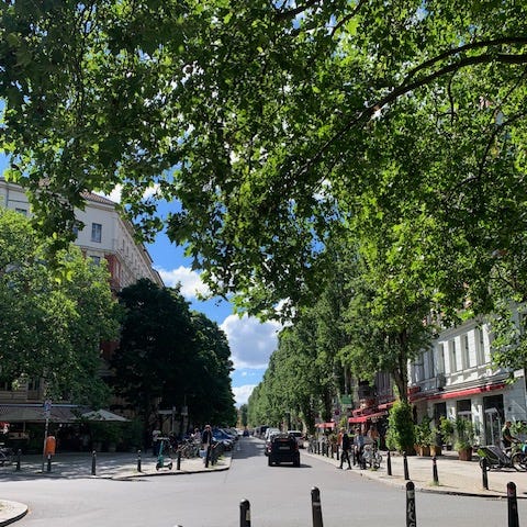 Photo of a treelined street in Berlin. It’s quiet with only few people and one car. The sky is a beautiful blue and the sun is shining.